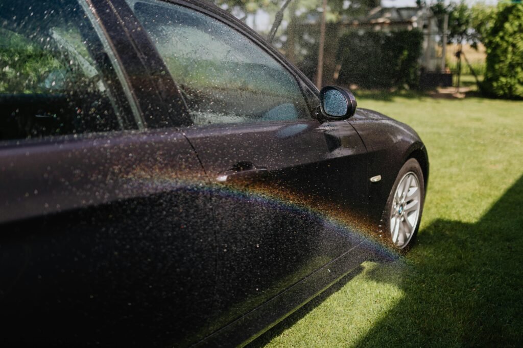 A black car being washed, capturing a rainbow in sunlight with water droplets on the exterior.