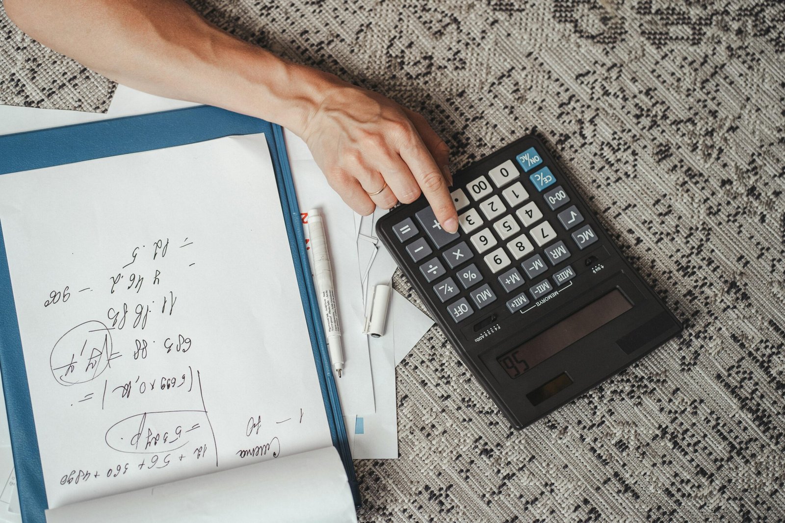 Top view of a hand calculating numbers with a large calculator and notes on a desk.